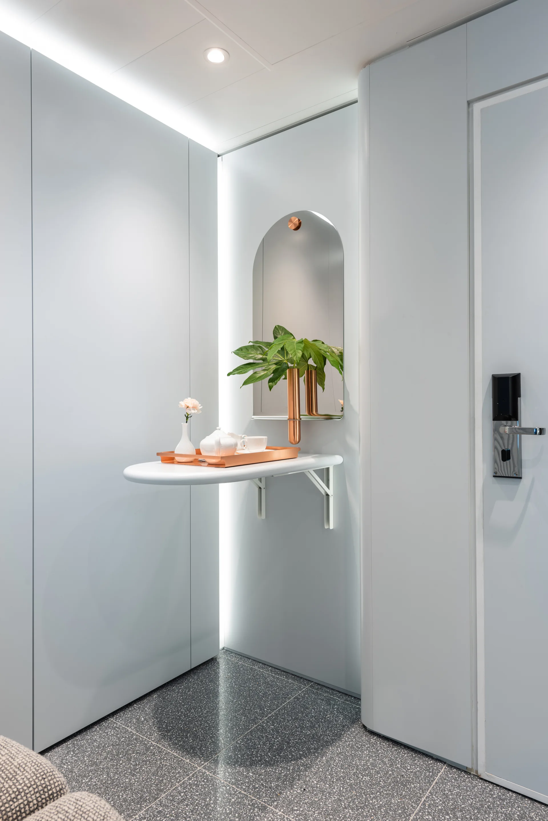 A small white wall-mounted console table stands in a light blue entryway niche beneath an arched mirror and a warm copper tray.