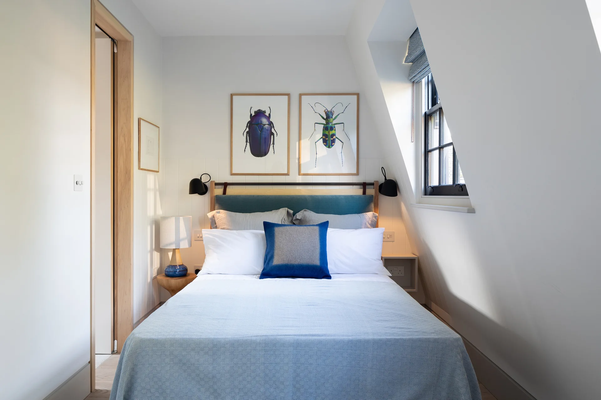 A compact attic bedroom features a bed with blue bedding, two framed beetle prints, and a window set into the sloped wall.