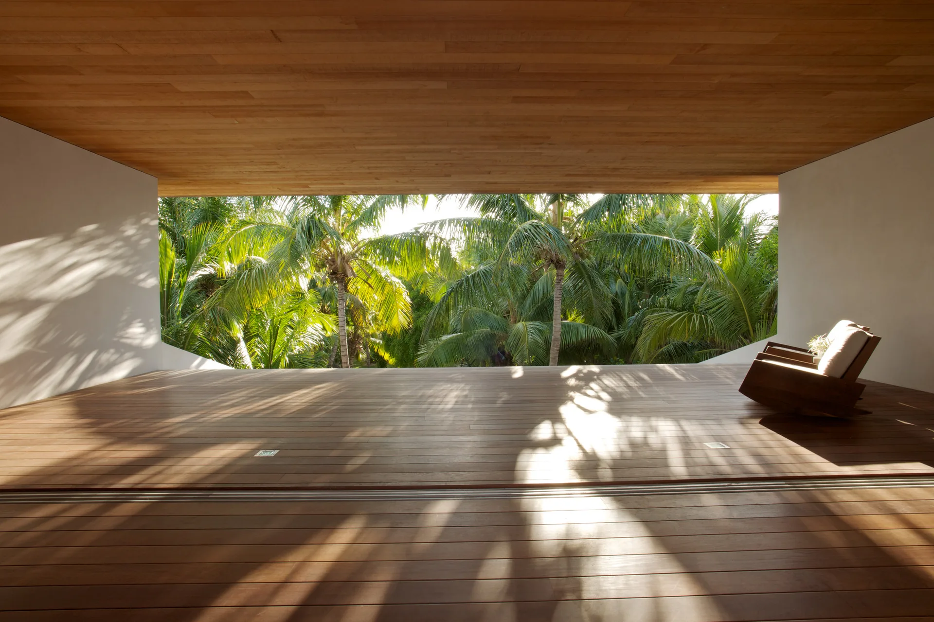 A covered wood deck with matching plank flooring and ceiling features a single lounge chair looking out at tropical palm trees.