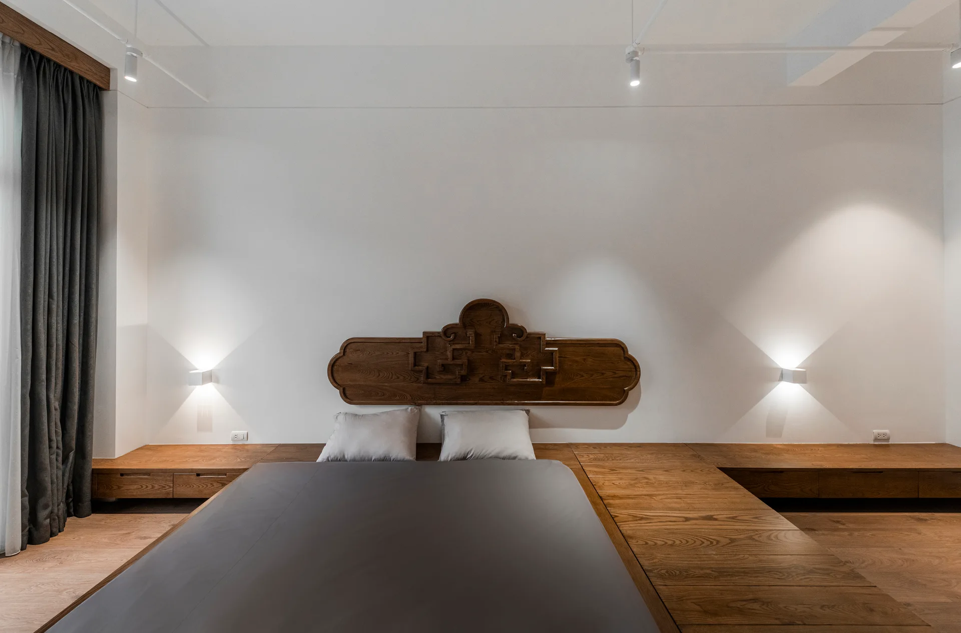 A minimalist bedroom featuring a low wooden platform bed with a carved wood headboard against white walls flanked by two glowing sconces.