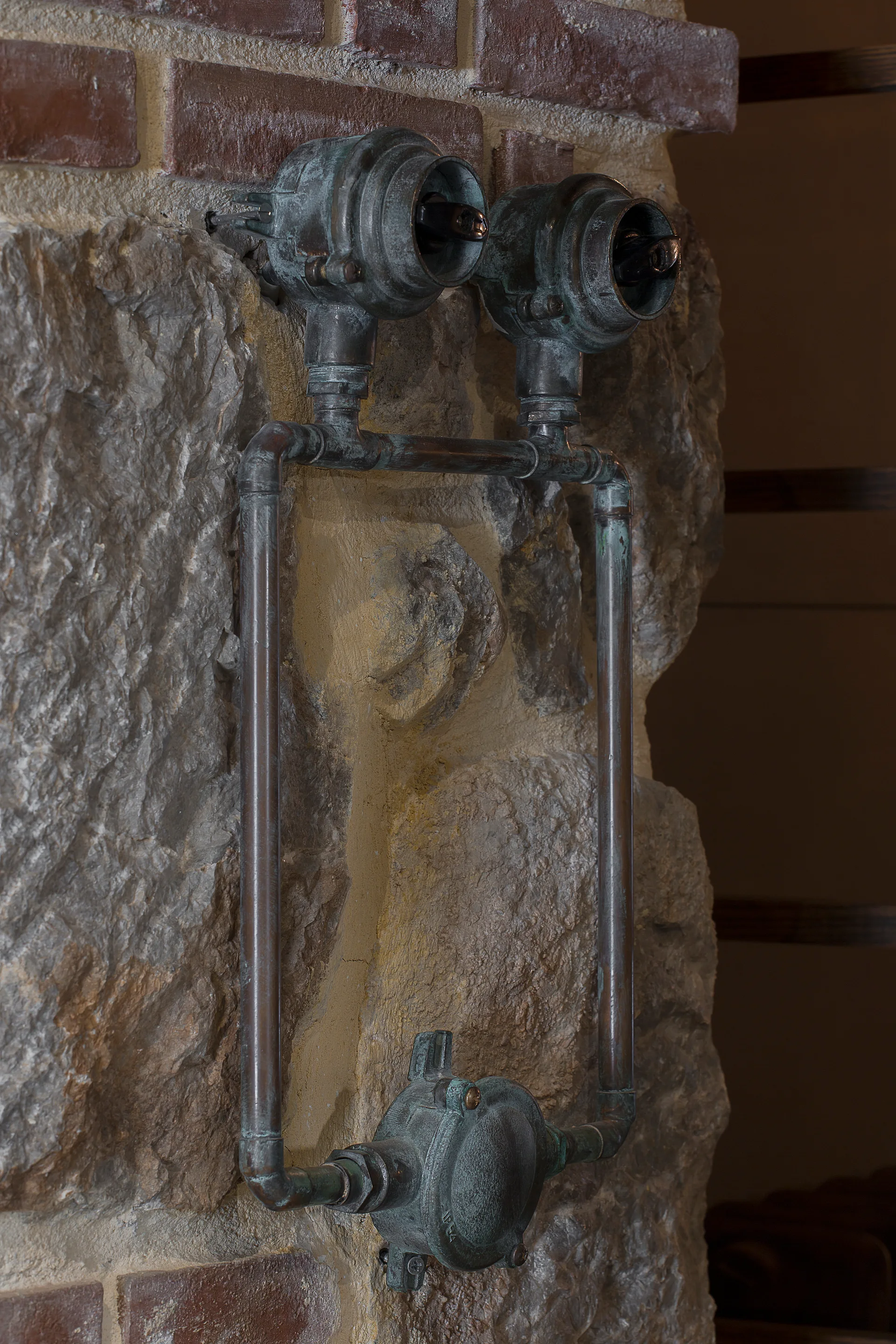 A close-up of patinated copper light switches and exposed metal conduits mounted on a textured stone and brick wall.