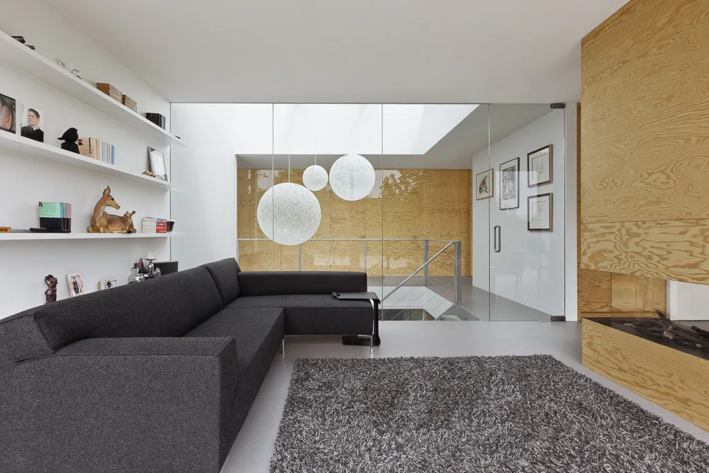 A modern living room features a dark grey sectional sofa, white floating shelves, and a plywood fireplace next to a large interior glass wall.