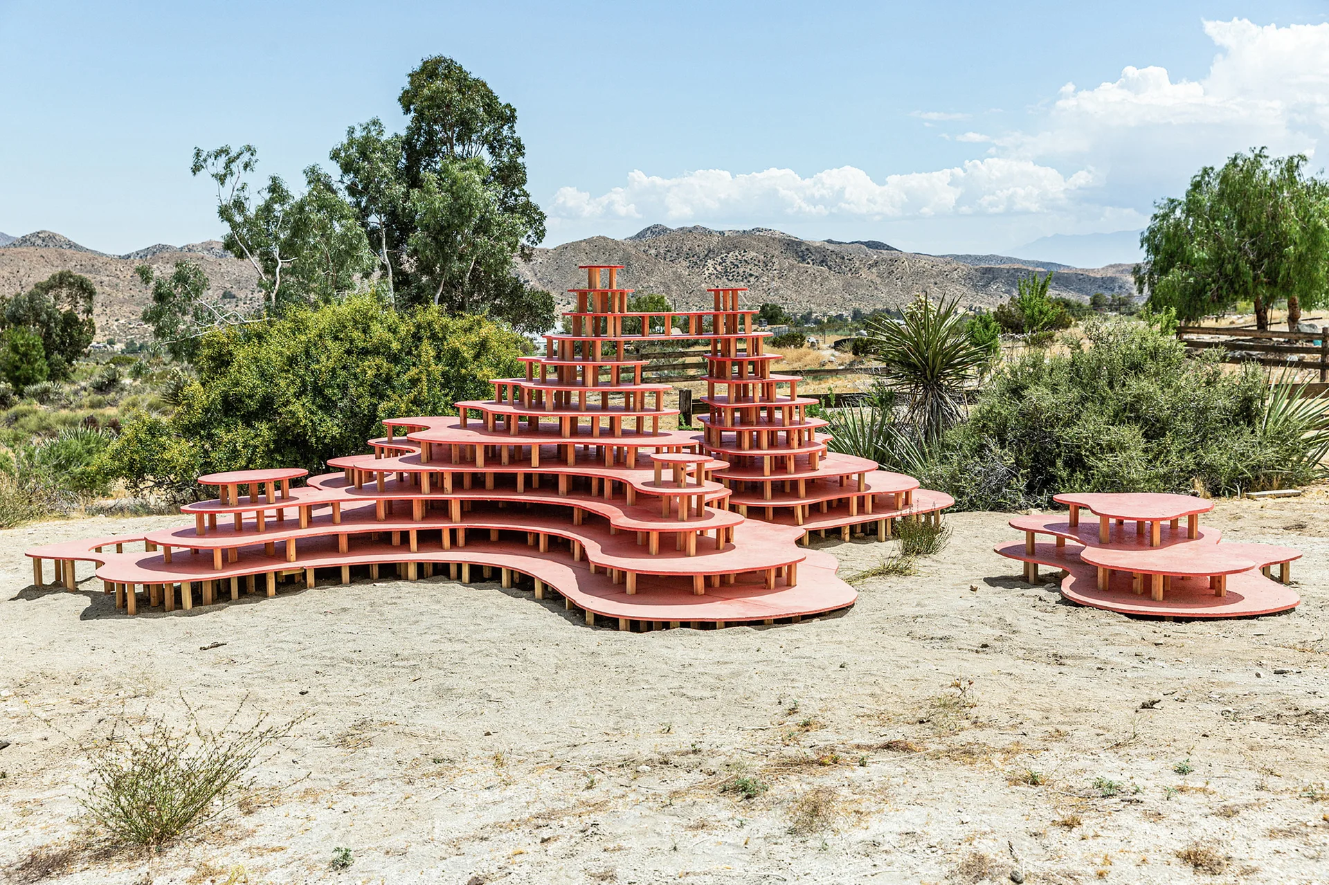 A multi-tiered wooden platform structure painted salmon pink stands on sandy desert ground with mountains and green shrubs in the background.