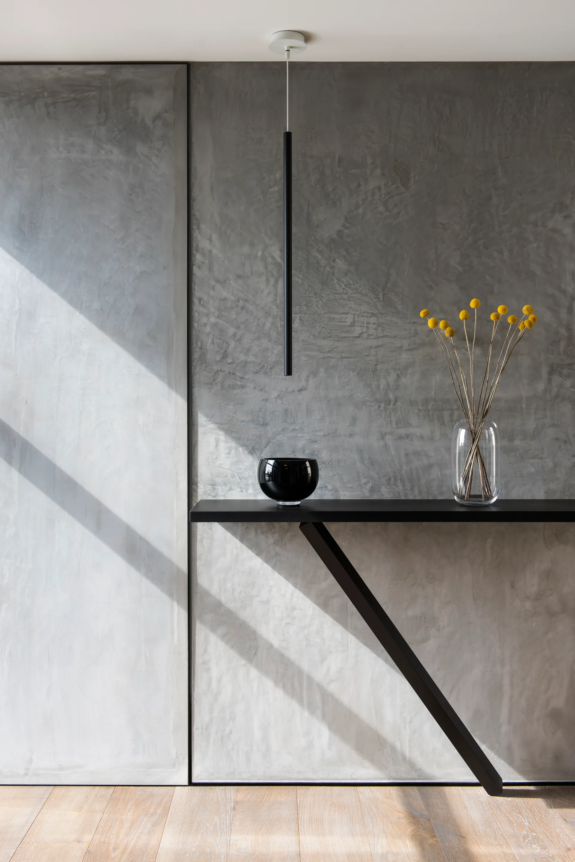 A minimalist black console table stands against a textured gray concrete wall with diagonal shadows and a clear vase of yellow flowers.