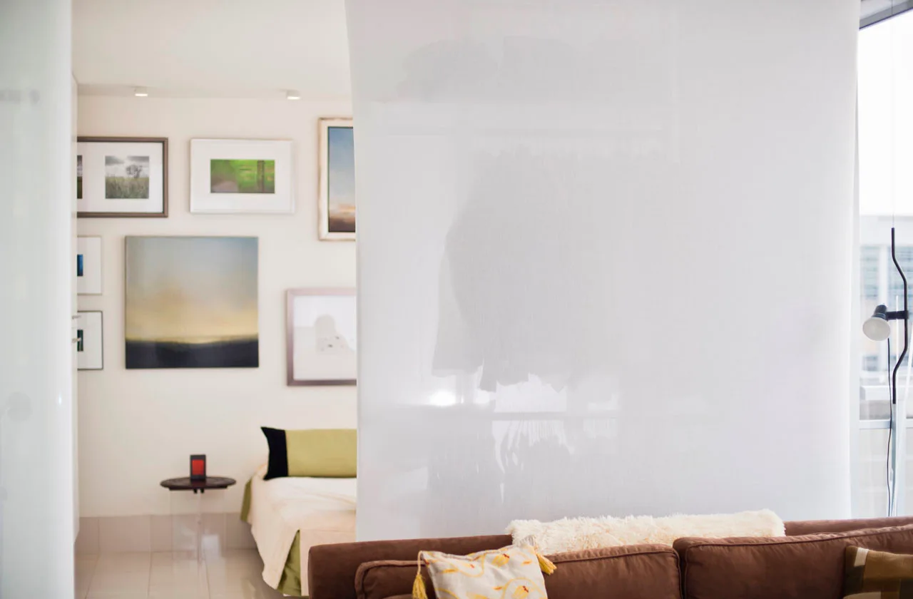 A translucent white partition separates a brown sofa in the foreground from a bedroom with a gallery wall in the background.