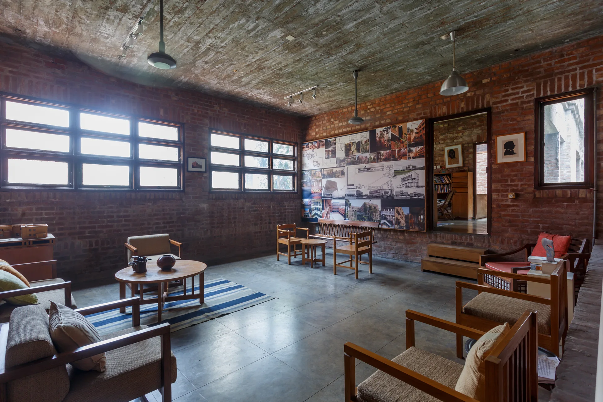 An industrial living room featuring exposed red brick walls, a raw concrete ceiling, wooden armchairs, and a large architectural photo collage.