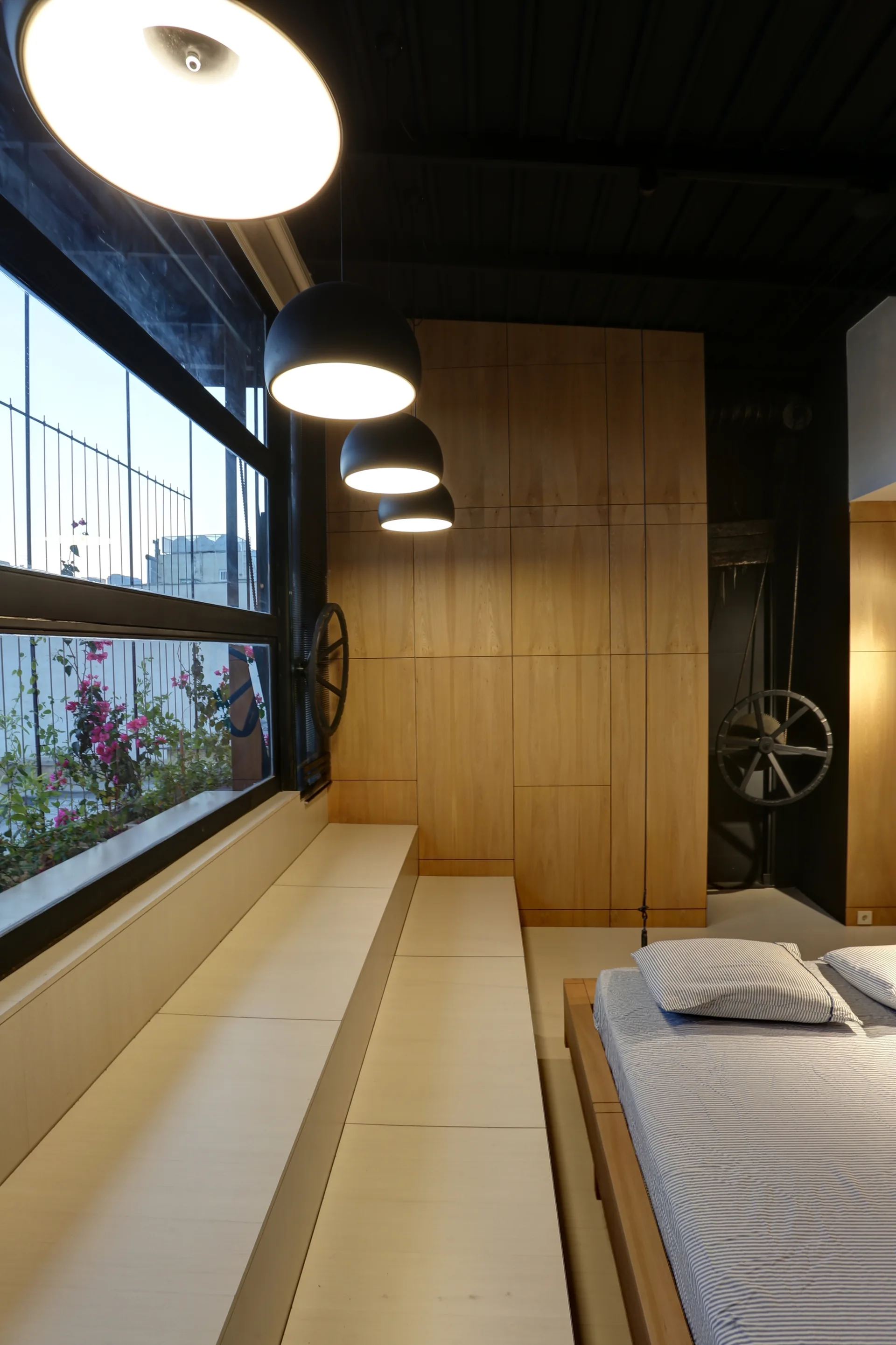A modern bedroom features a low platform bed with striped sheets, a stepped window bench, black pendant lights, and a light wood paneled wall.