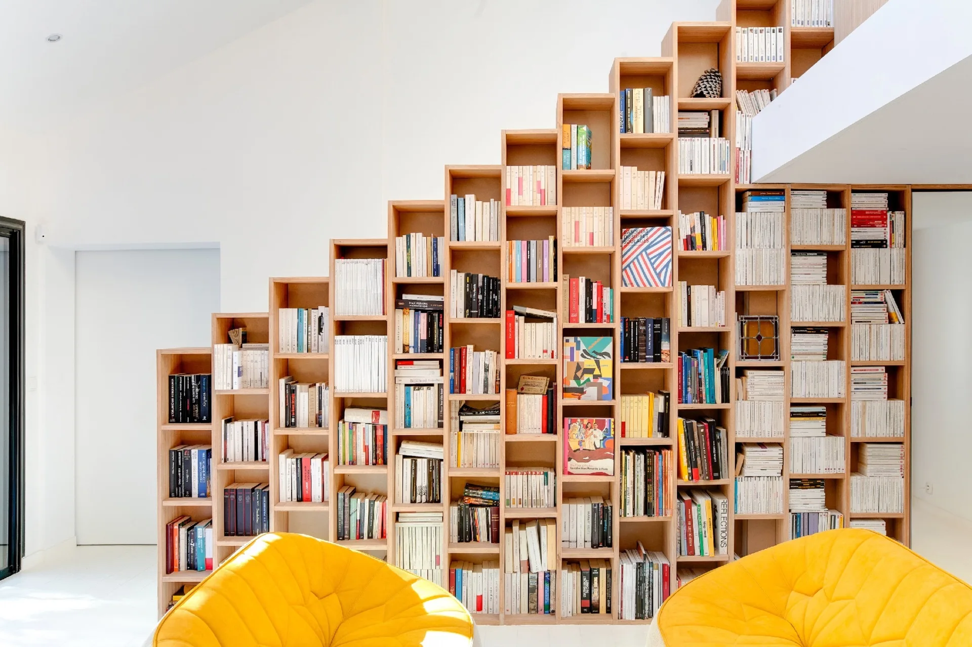 A tall stepped wooden bookcase filled with books stands against a white wall behind two tufted yellow armchairs.