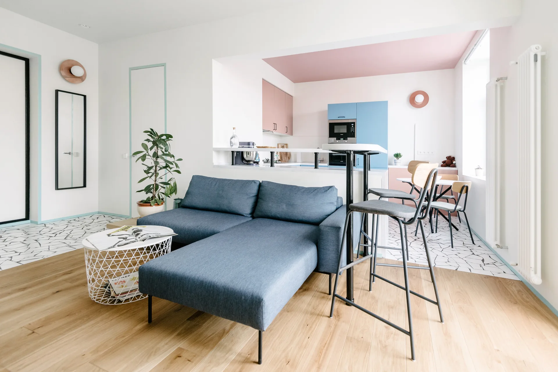 A contemporary living room featuring a blue chaise sofa and wood flooring transitioning into a kitchen with pink and blue cabinets and geometric tile.