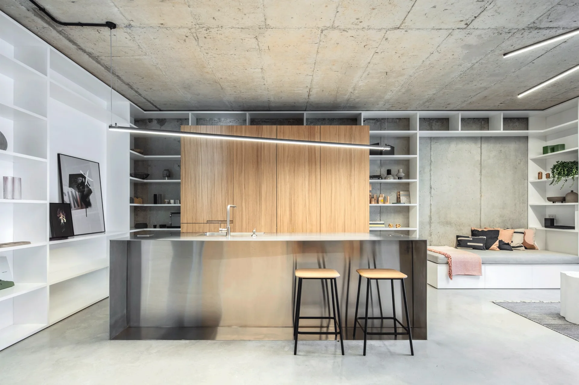 A modern industrial kitchen with a stainless steel island, two wooden bar stools, built-in white shelving, wood cabinetry, and an exposed concrete ceiling.