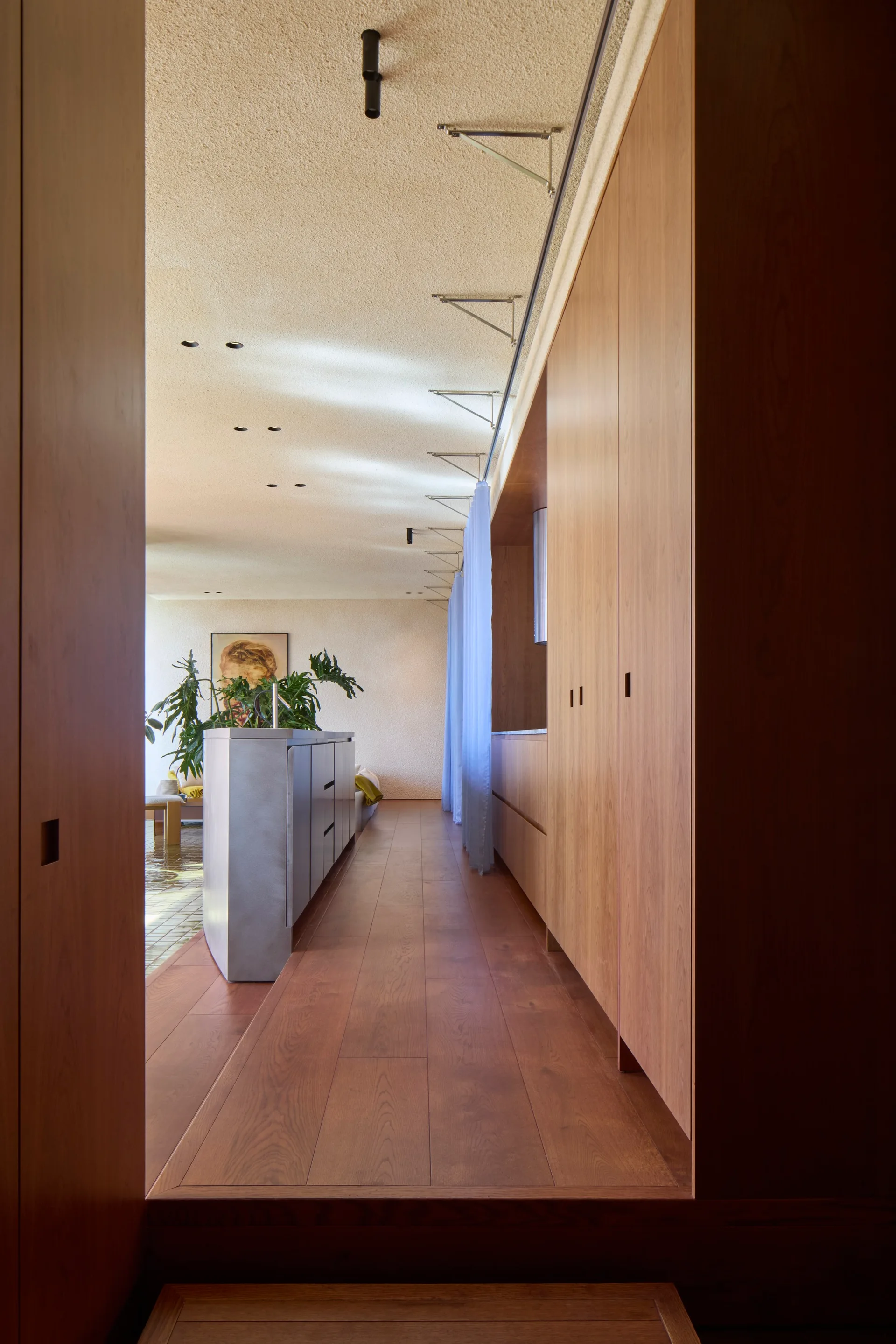 A hallway view of a minimalist kitchen featuring floor-to-ceiling wood cabinetry, a sleek metallic island, and sheer blue curtains.