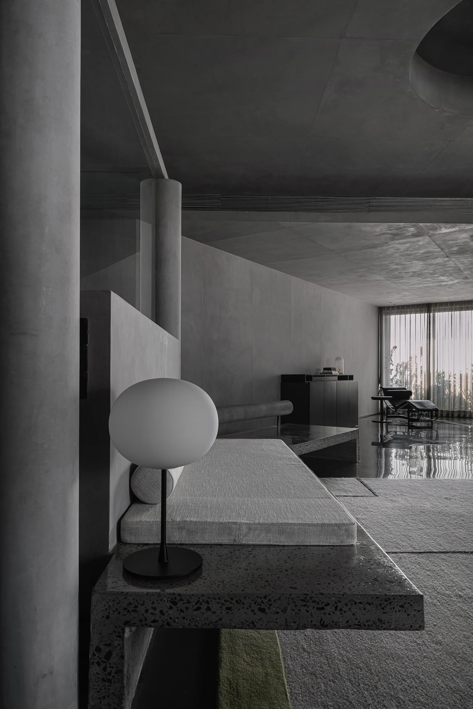 A minimalist grey living room features a built-in terrazzo platform daybed with a globe table lamp next to a concrete column and large windows.