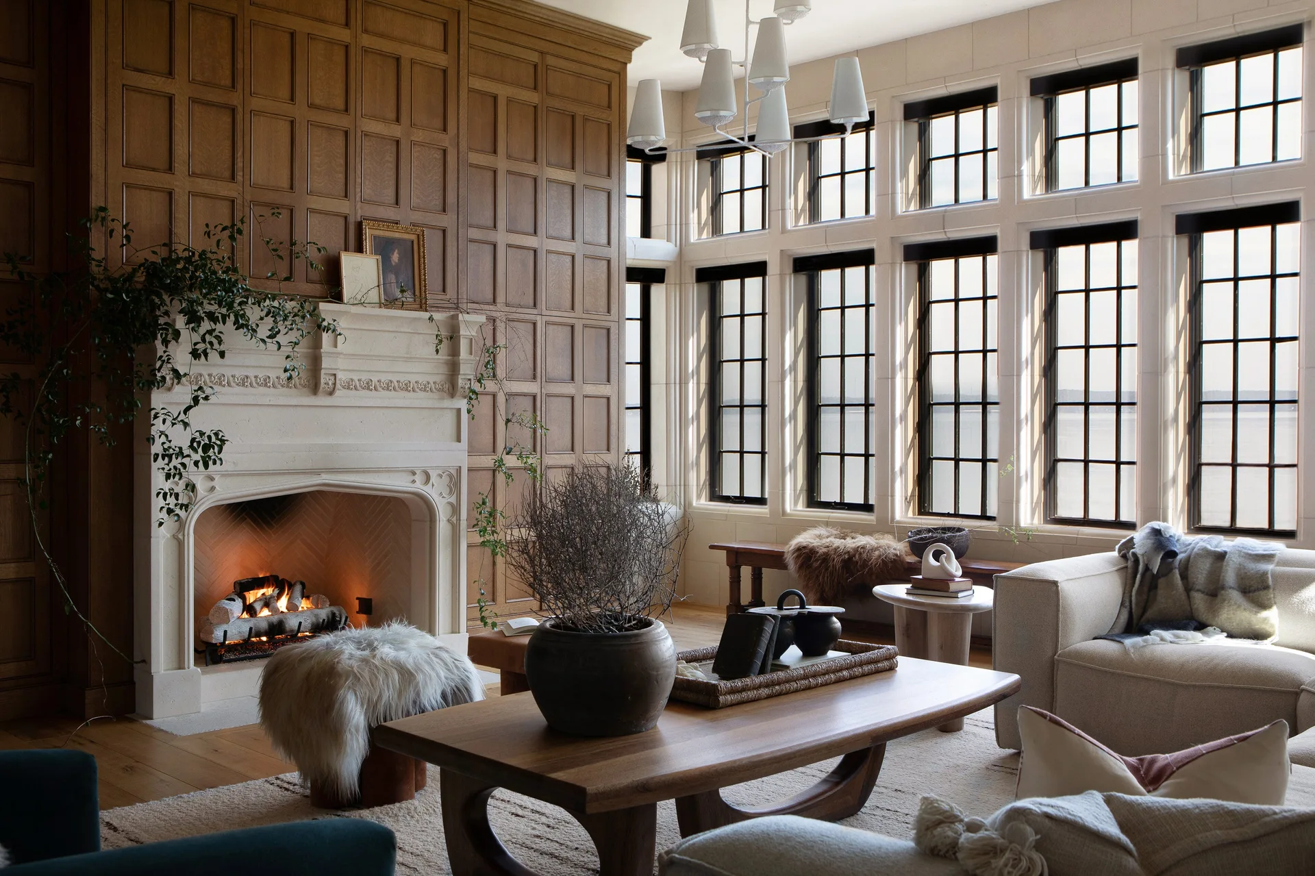 A living room features a carved stone fireplace set against wood paneling next to large grid windows overlooking water.
