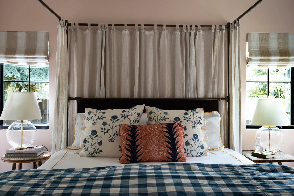 A symmetrical bedroom features a black metal canopy bed with linen drapes, floral pillows, a blue gingham quilt, and glass lamps on woven side tables.