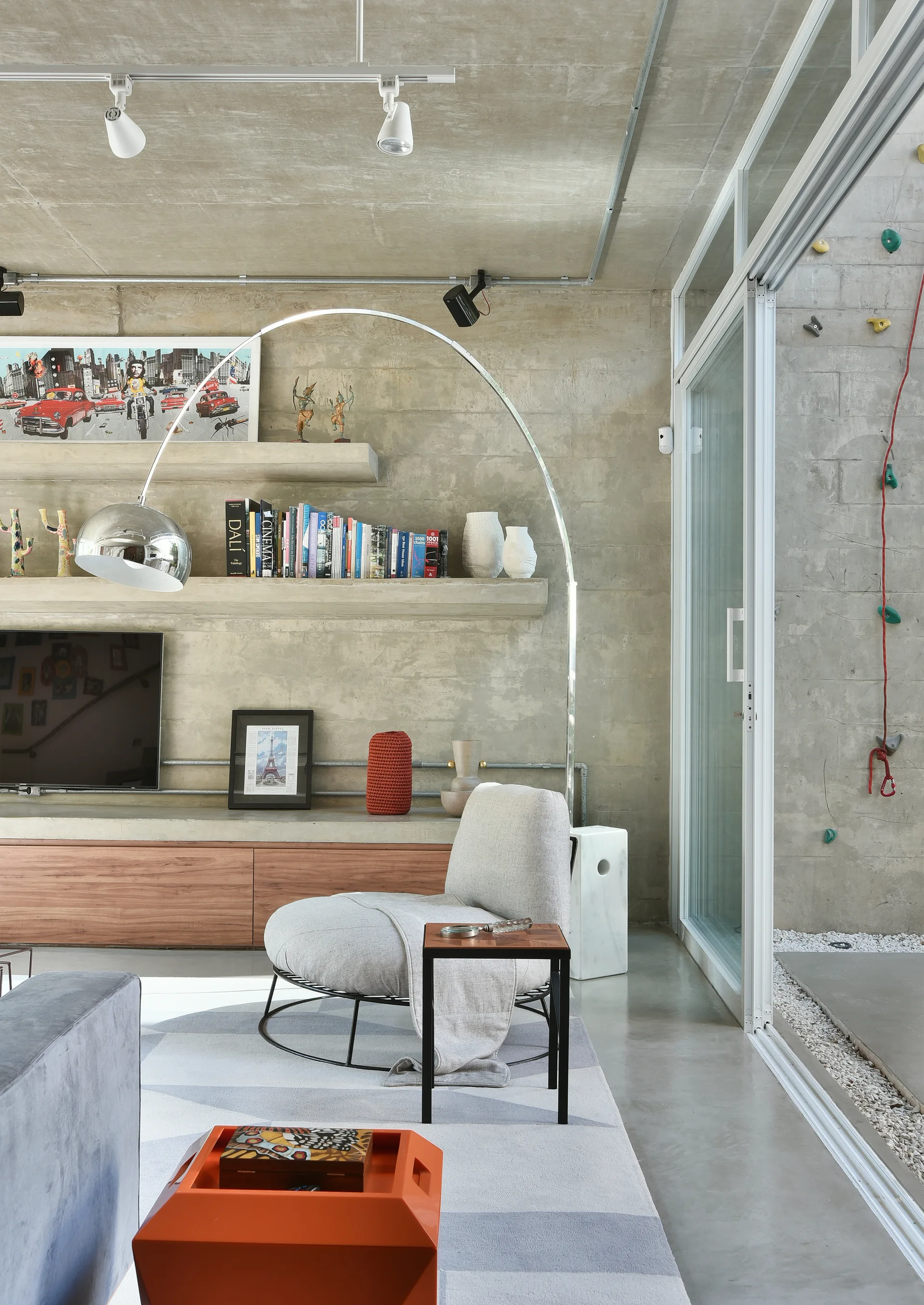 A modern living room with an exposed concrete wall, floating shelves, an armchair under an arch lamp, and a climbing wall behind a sliding glass door.