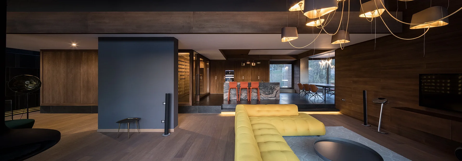 A modern open-plan living room features a large yellow tufted sofa on a grey rug, set against dark wood walls and a kitchen with a marble island.