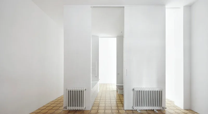 A minimalist white interior features a patterned tiled floor, two wall-mounted radiators, and a sliding pocket door leading to a bathroom.