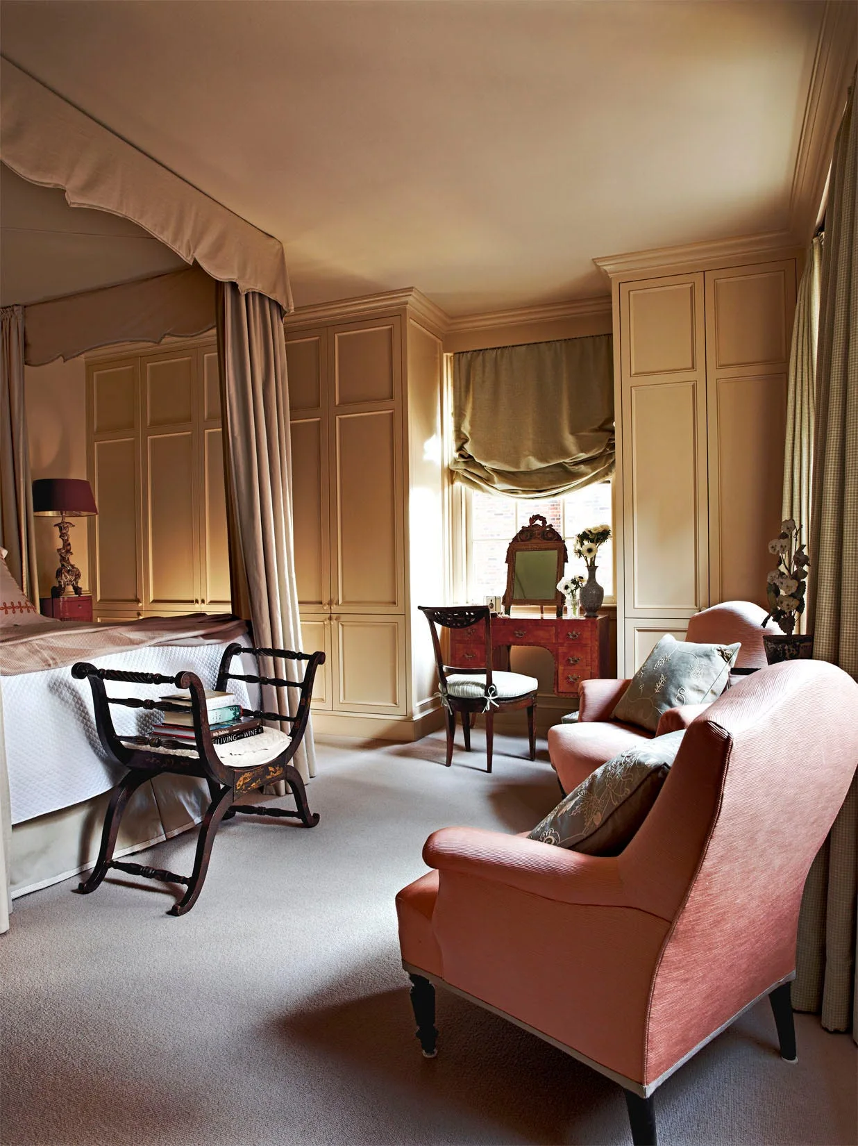 A traditional cream bedroom features a canopy bed, floor-to-ceiling built-in wardrobes framing a vanity table by the window, and two peach armchairs.