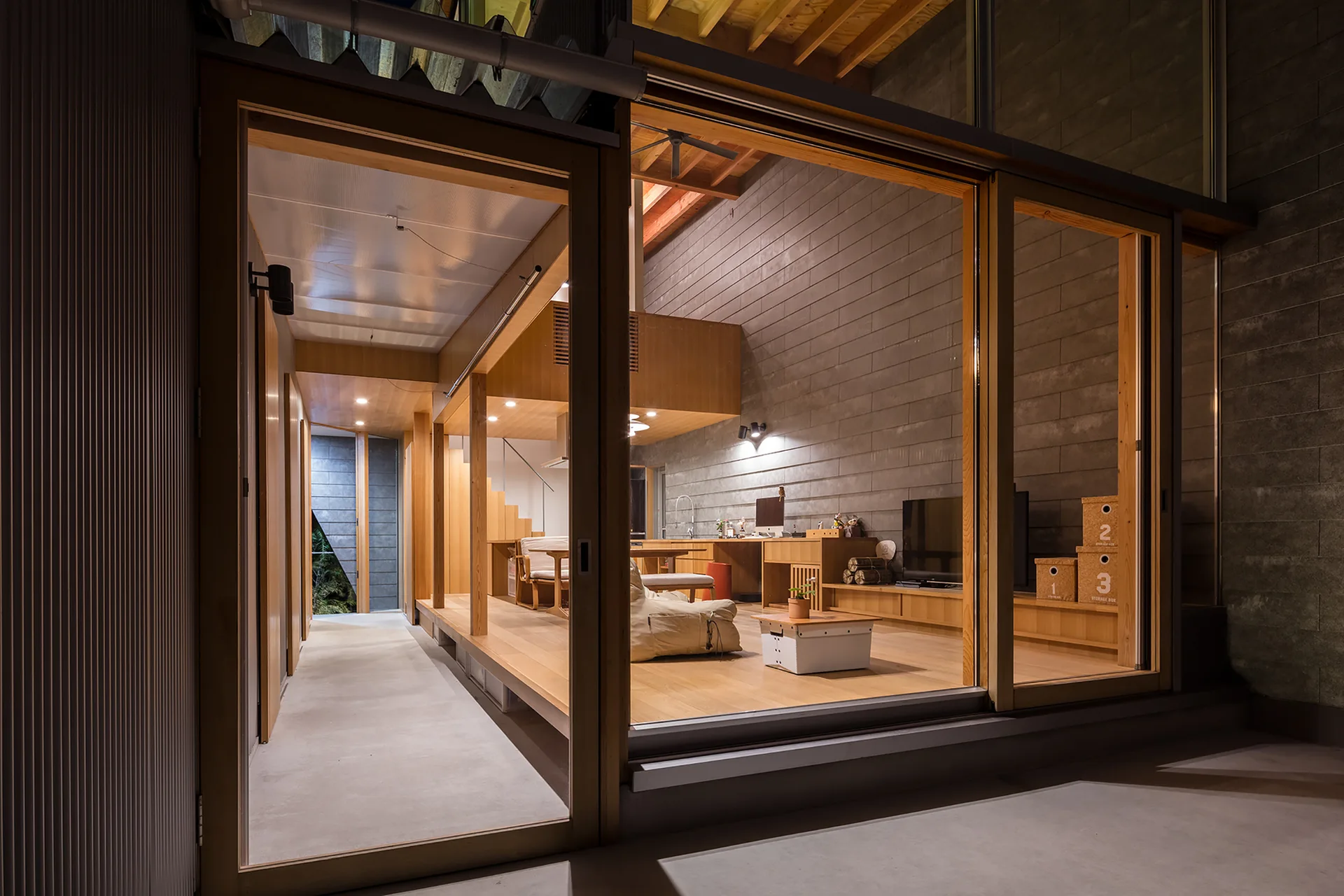 A modern open-plan living room with warm wood flooring and concrete block walls is seen through large sliding glass doors.