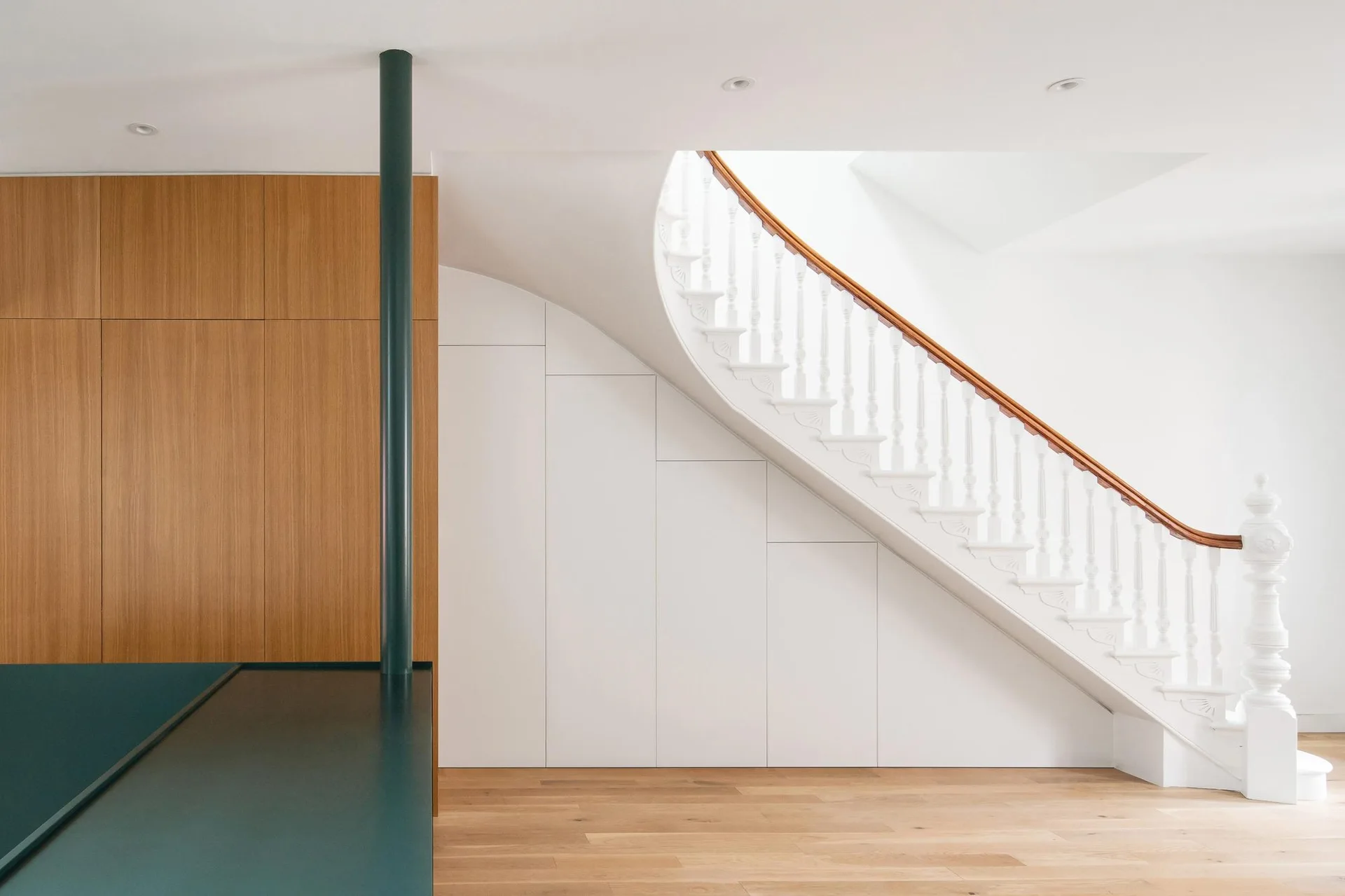 A curved white staircase with a wooden handrail rises above seamless built-in under-stairs storage cabinets and light wood floors.