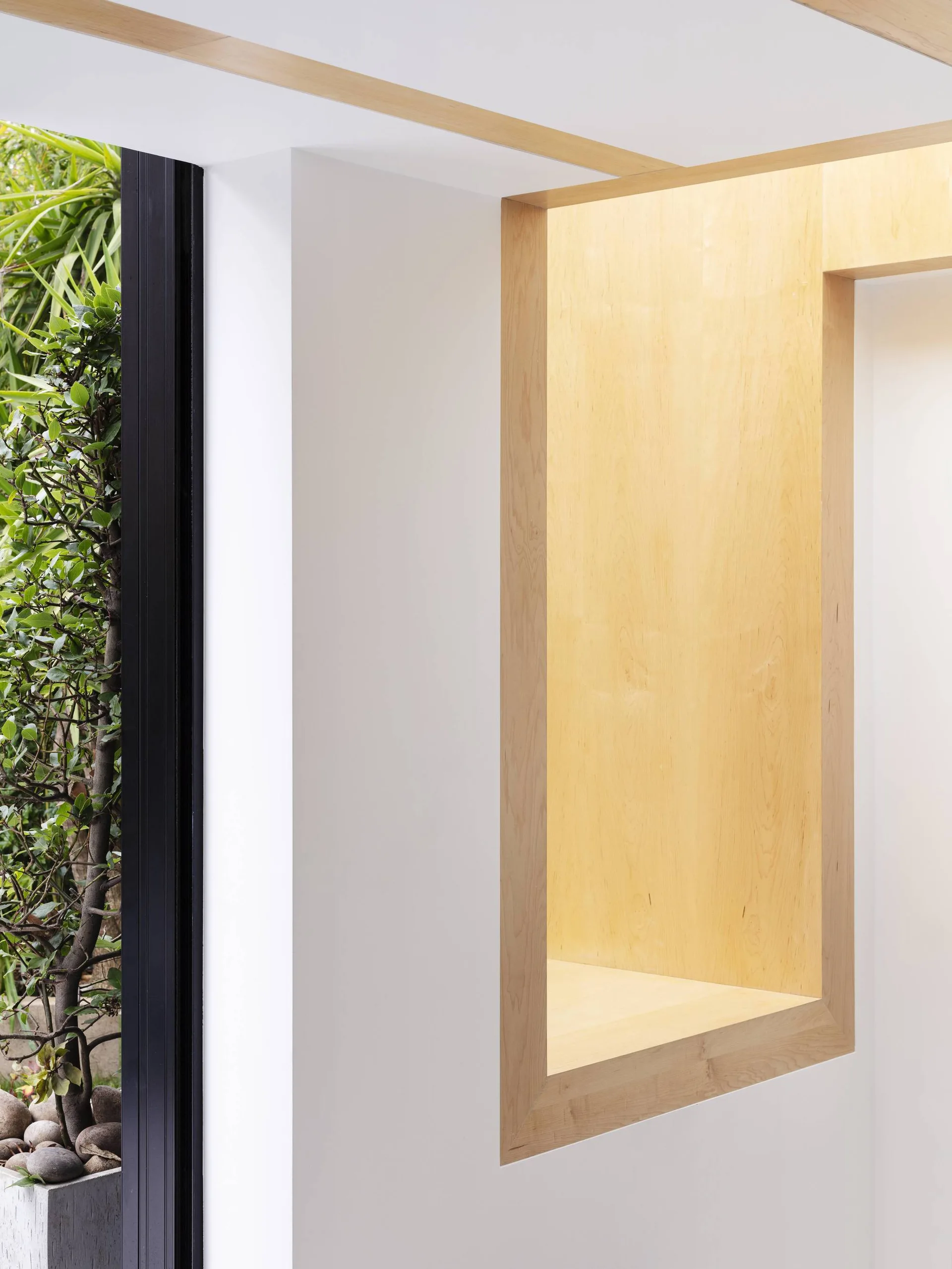A recessed light wood alcove set into a white wall next to a black-framed window showing green plants outside.