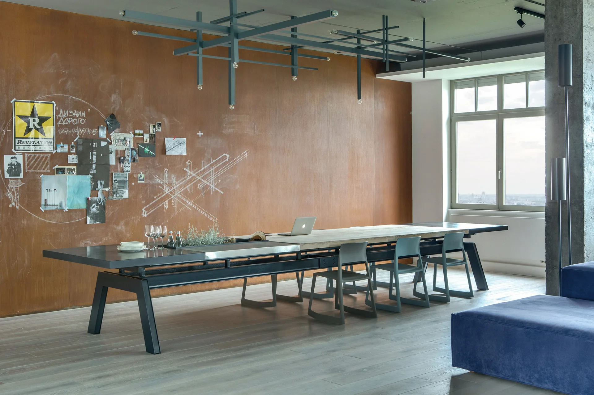 A long industrial dining table sits in front of a textured rust-colored accent wall beneath a dark gray geometric grid ceiling light.