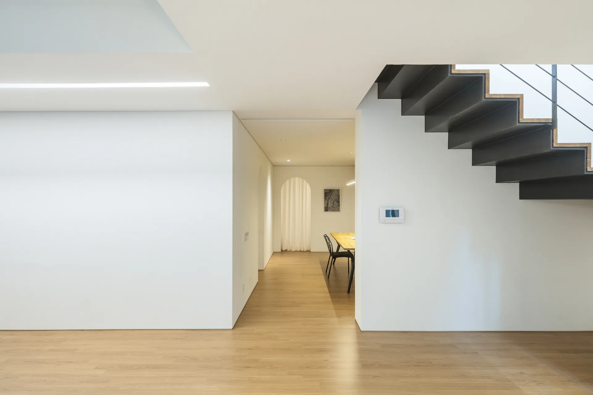 A modern minimalist hallway with white walls, light wood floors, an open-tread black staircase, and an arched door in the background.