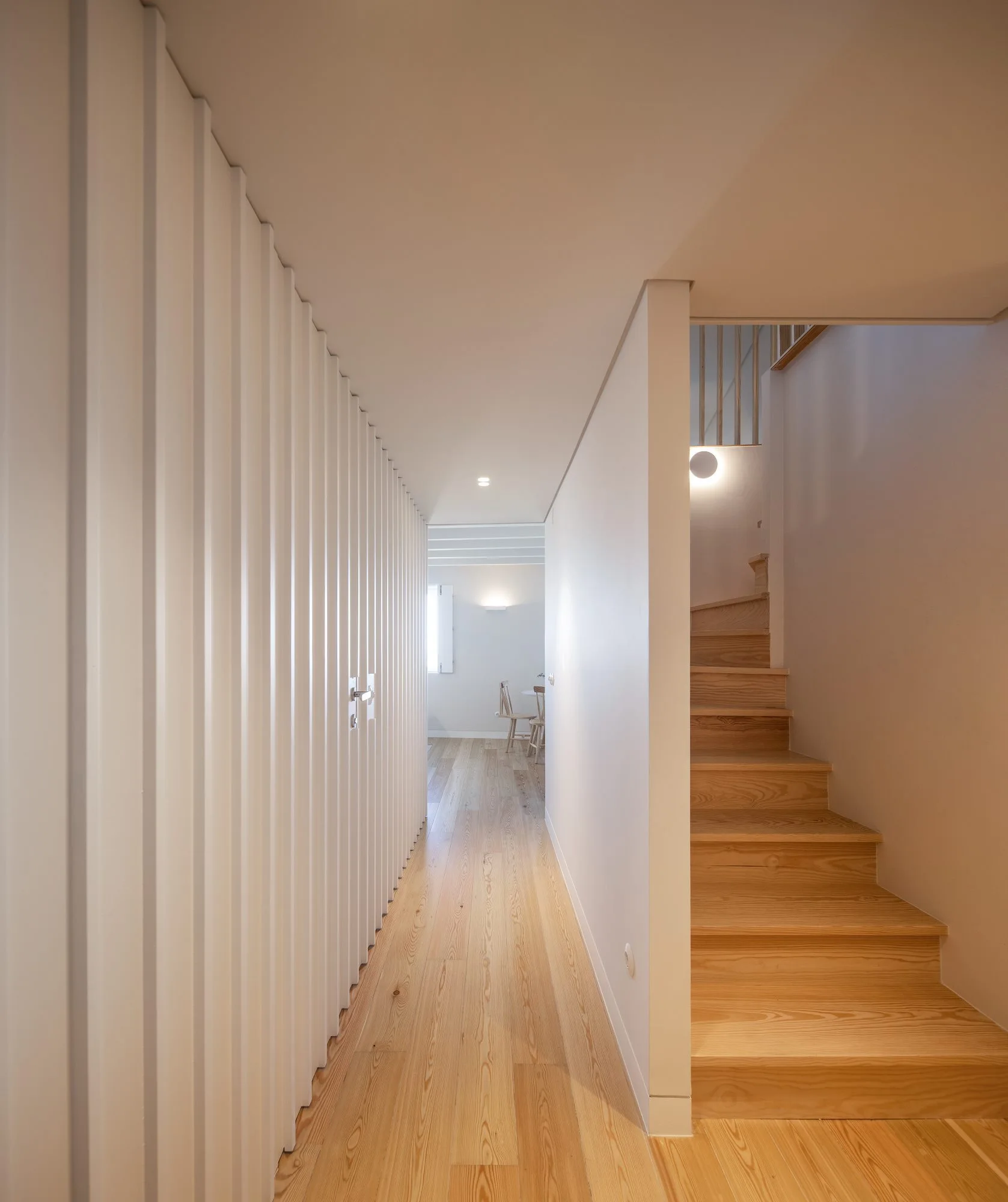 A minimalist hallway features light wood stairs on the right and a textured white vertical slatted wall lining the left side.