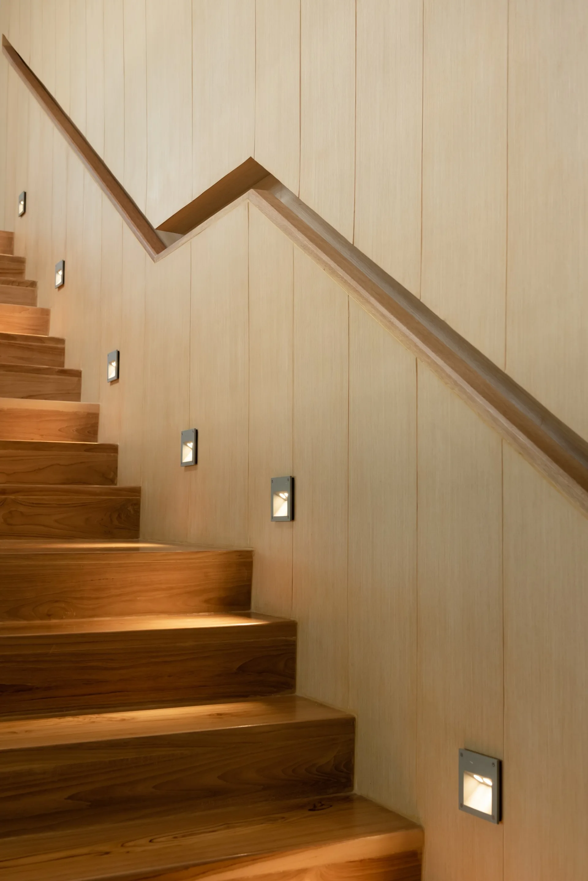 A wooden staircase ascends next to a wall with vertical light-wood paneling and square recessed light fixtures illuminating the steps.