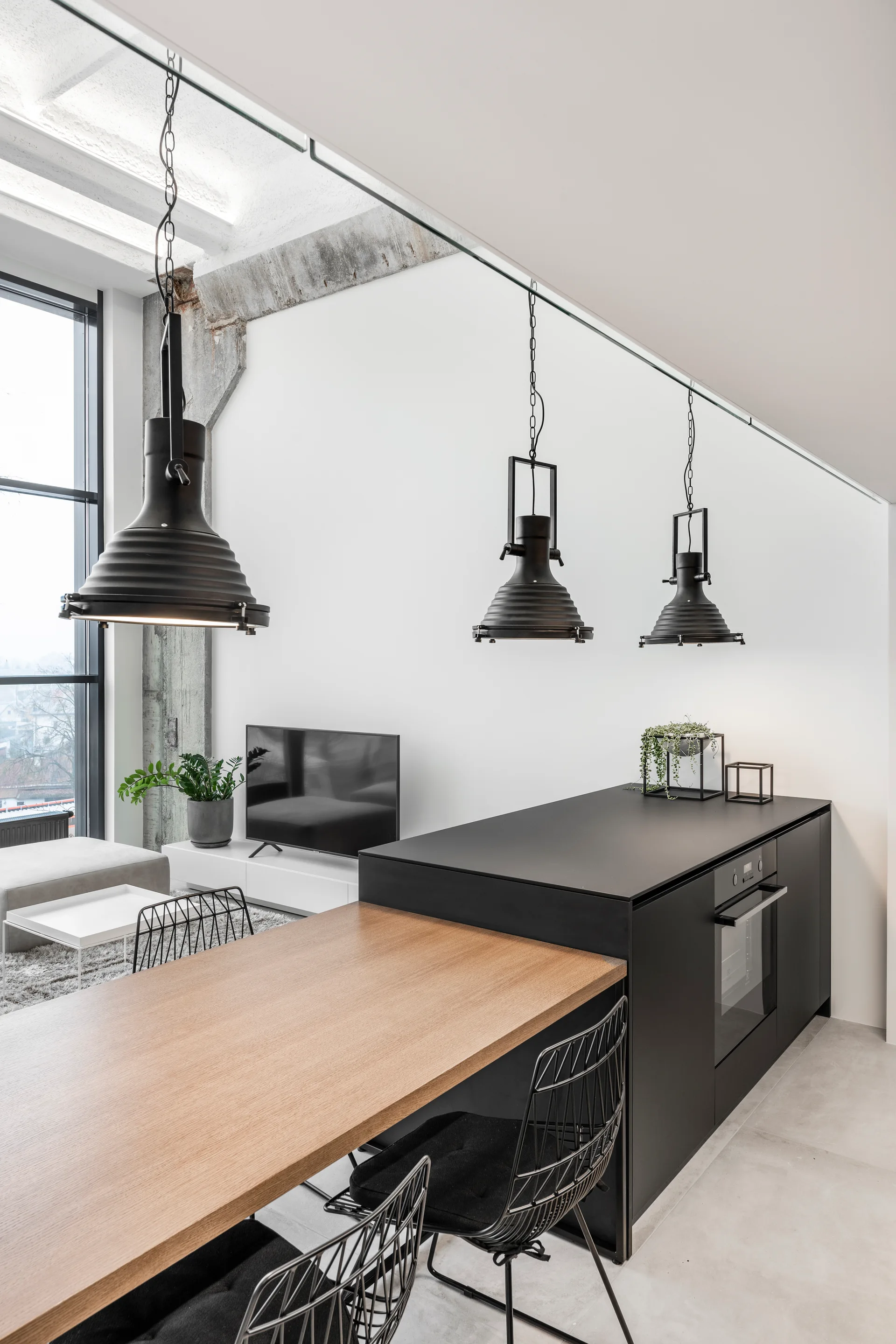 A modern kitchen features a matte black island with an integrated oven, a light wood table extension, and three industrial pendant lights.