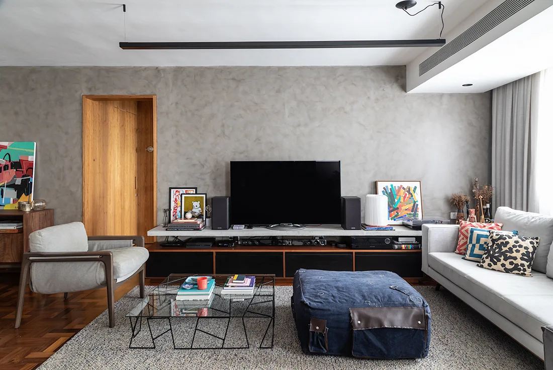 A contemporary living room with a textured grey plaster wall, a low wooden media console, a light grey sofa, and a large blue fabric ottoman.