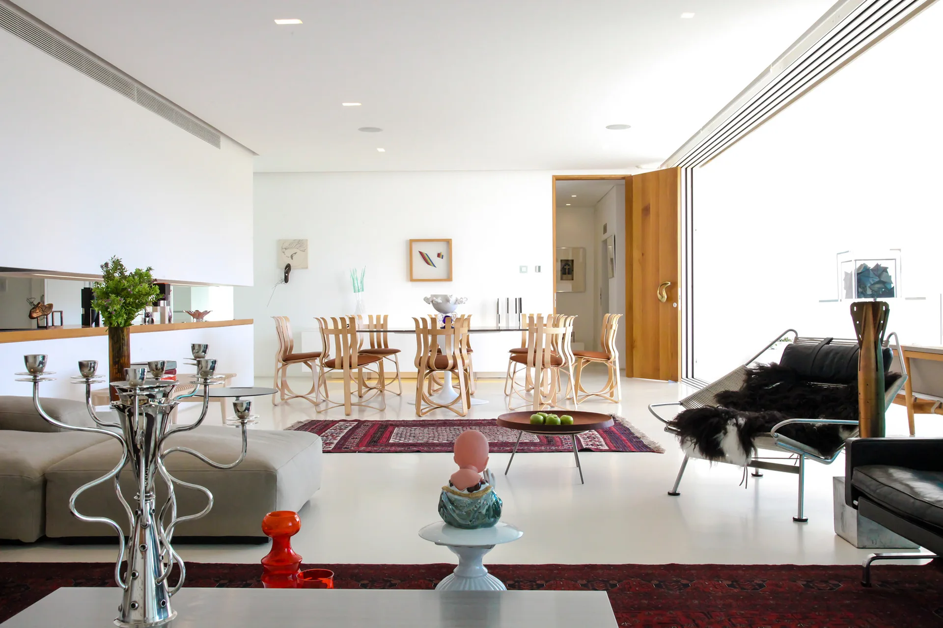 A bright open-concept living and dining room features white floors, curved wooden dining chairs, a gray sectional sofa, and patterned rugs.