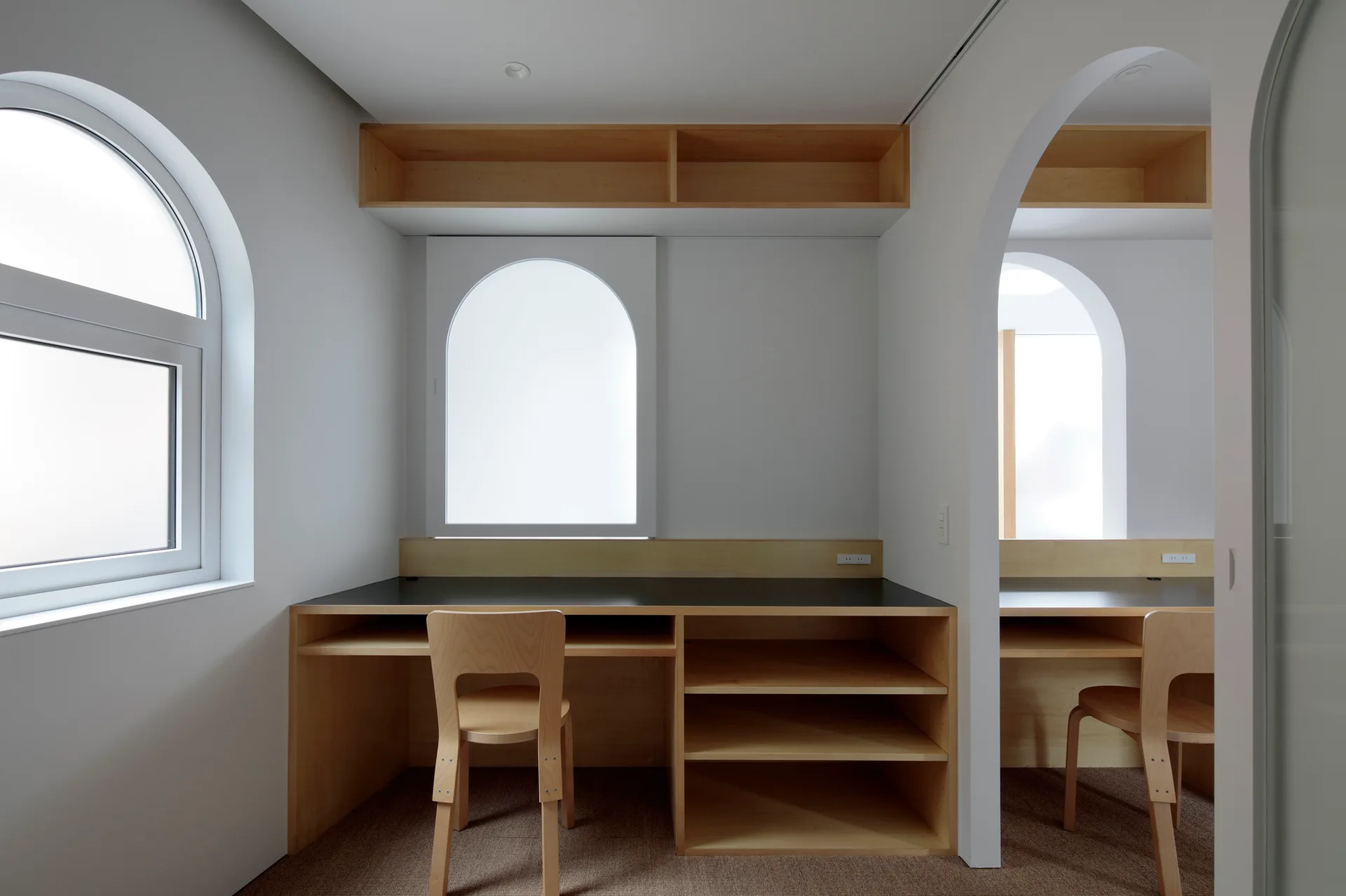 A minimalist home office featuring built-in light wood desks with black tops, matching chairs, and white walls with arched windows and doorways.