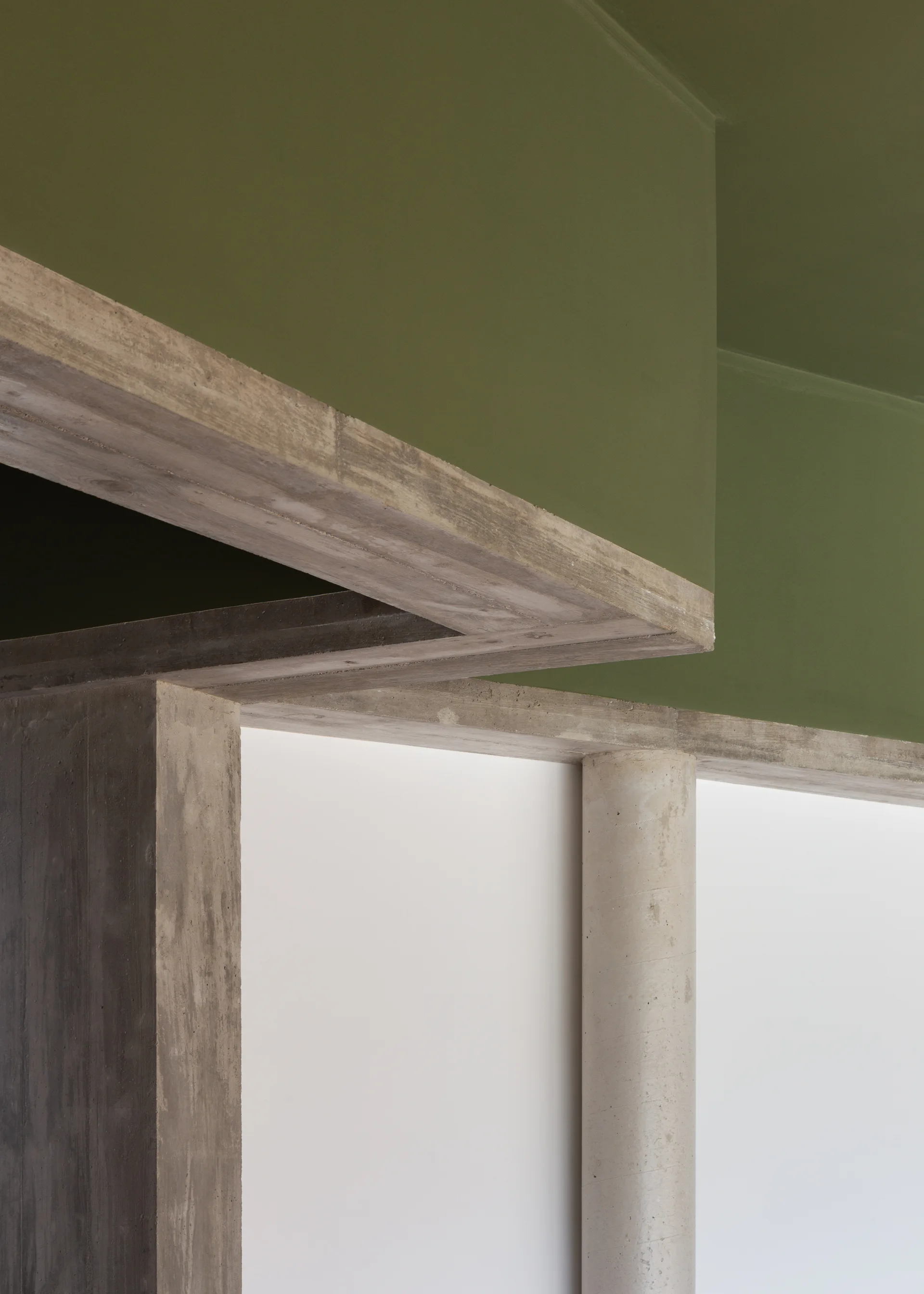 Architectural detail of a cylindrical concrete column and raw concrete beams set against a white wall and olive green painted ceiling.