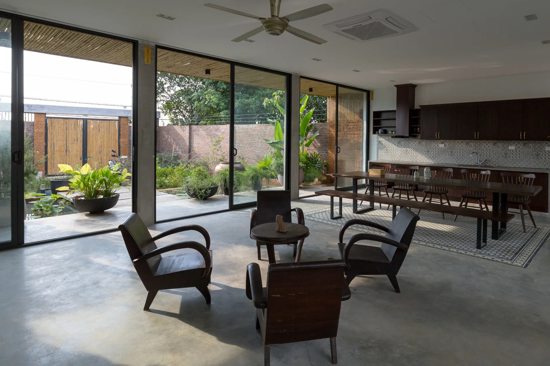 An open-plan living space with gray concrete floors, four dark wooden armchairs, and large sliding glass doors opening to a green outdoor garden.
