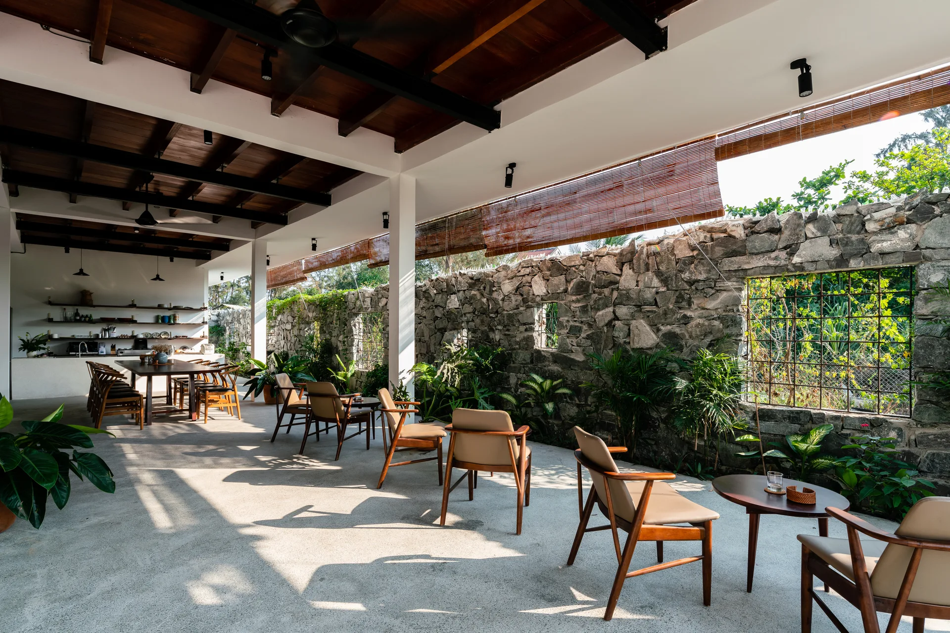 Mid-century wooden armchairs sit on a concrete patio beside a weathered stone boundary wall beneath an exposed beam ceiling.