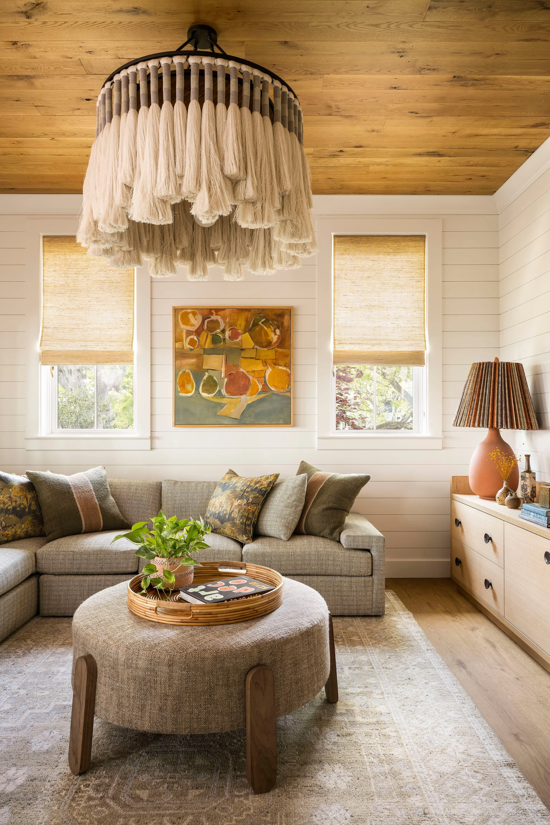 A living room features a large tassel chandelier hanging from a wood plank ceiling, flanked by two windows over a grey sofa with an ottoman.