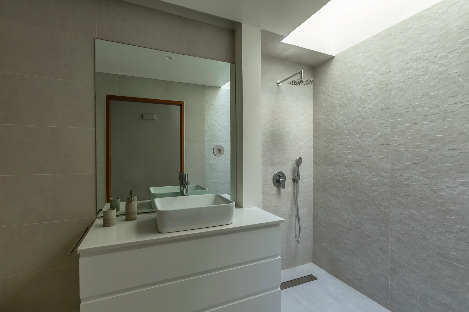 A minimalist bathroom features a white vanity with a rectangular vessel sink next to a walk-in shower beneath a bright skylight.