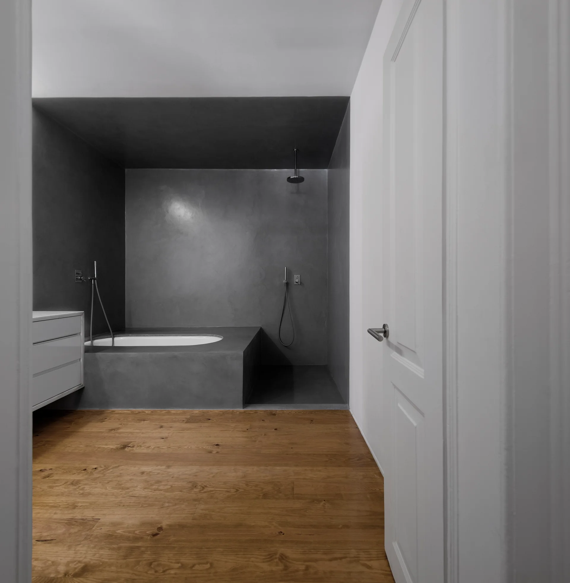 A minimalist bathroom features a dark grey recessed tub and shower area contrasted by warm wood floor planks and a white vanity cabinet.