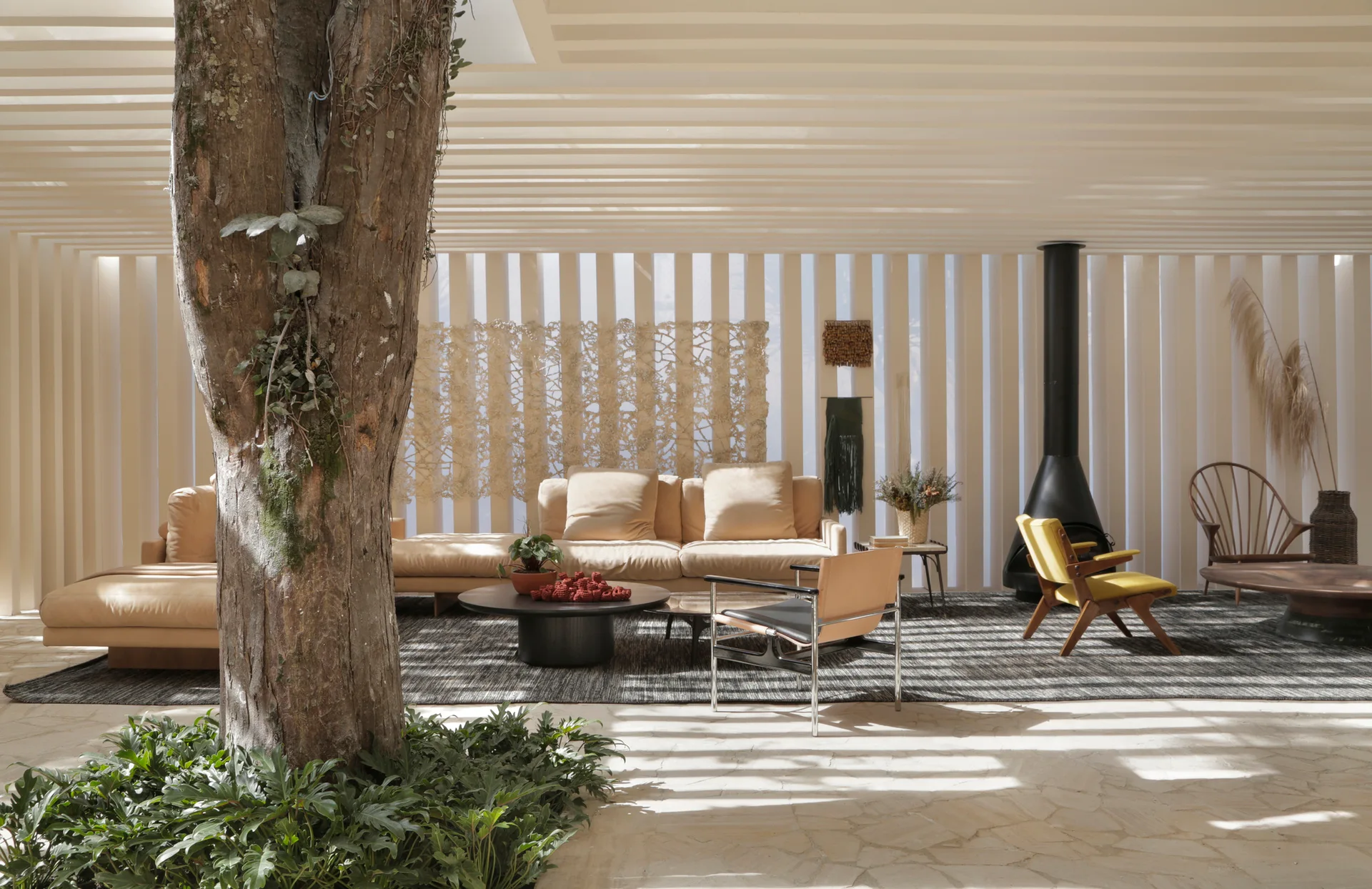 An organic modern living room featuring an integrated indoor tree trunk, slatted walls, and a tan sectional sofa on a stone tile floor.