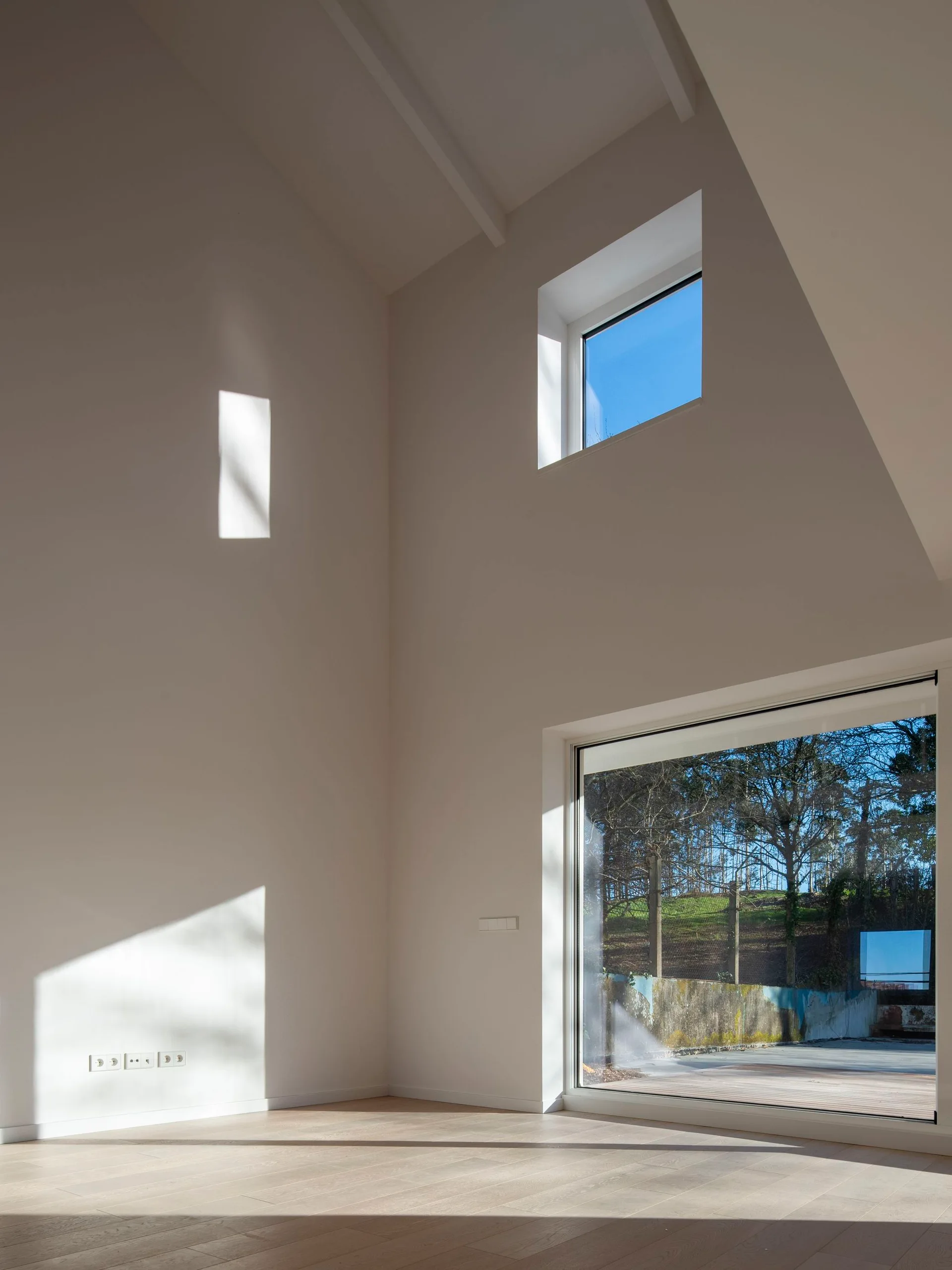 An empty minimalist room features high vaulted white ceilings, light wood floors, and sunlight streaming through two different sized square windows.