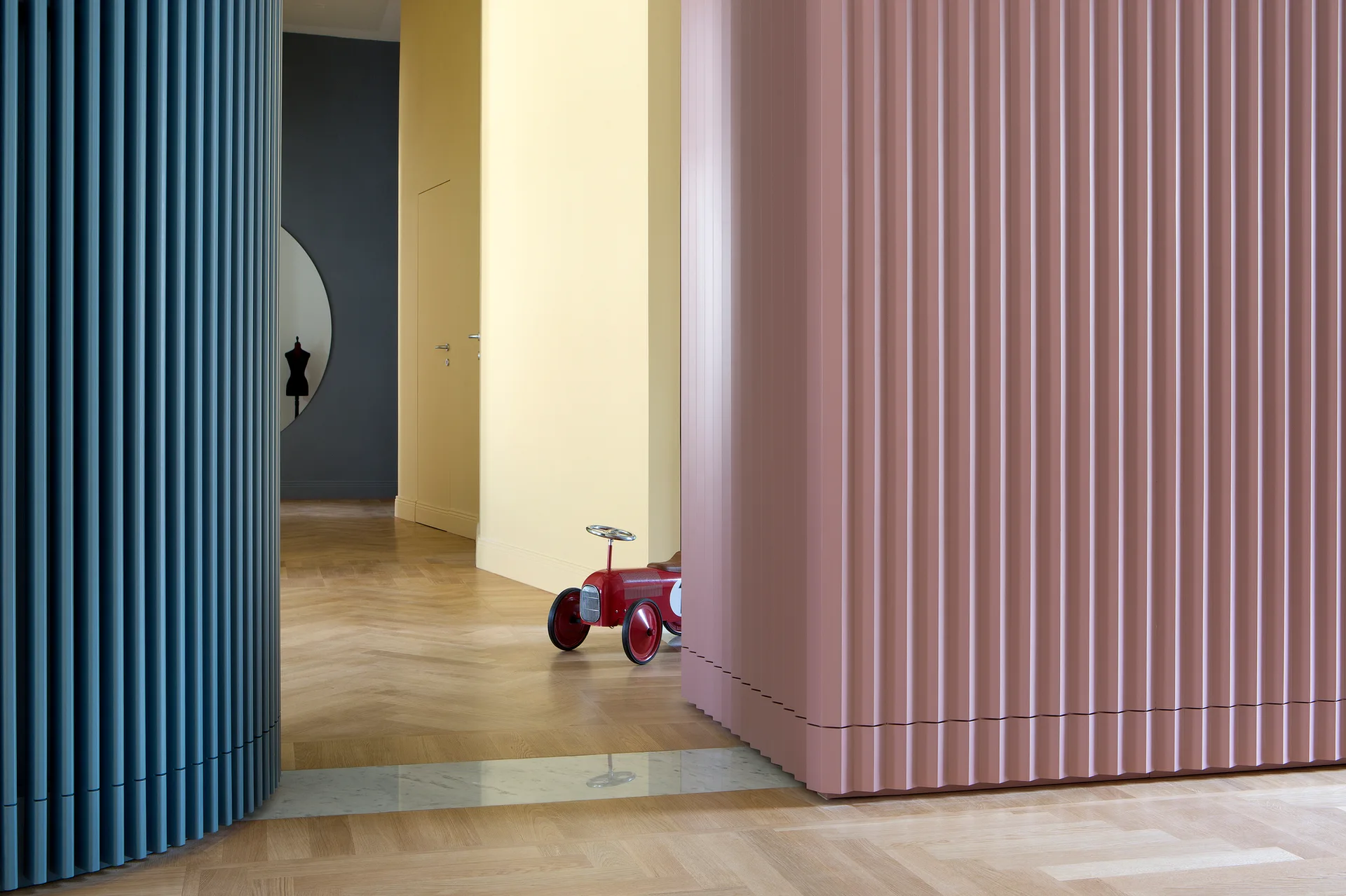 A modern residential hallway features curved fluted wall panels in blue and dusty pink, light wood herringbone flooring, and a yellow background wall.