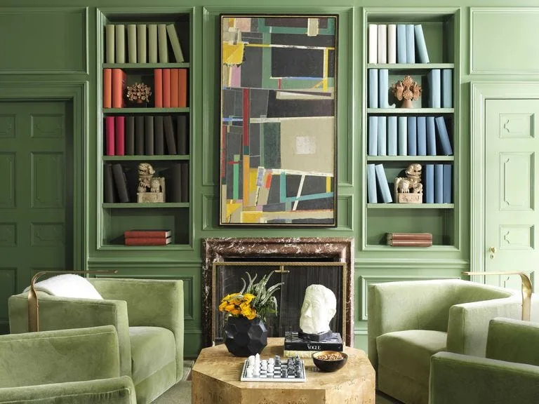 A symmetrical green living room features velvet armchairs, built-in bookshelves flanking a marble fireplace, and an abstract painting.