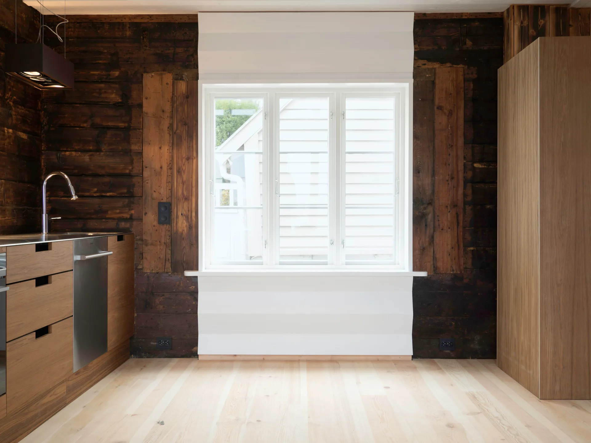 A modern rustic kitchen features dark brown timber plank walls, a central white window frame, and light wood cabinetry with a metal faucet.
