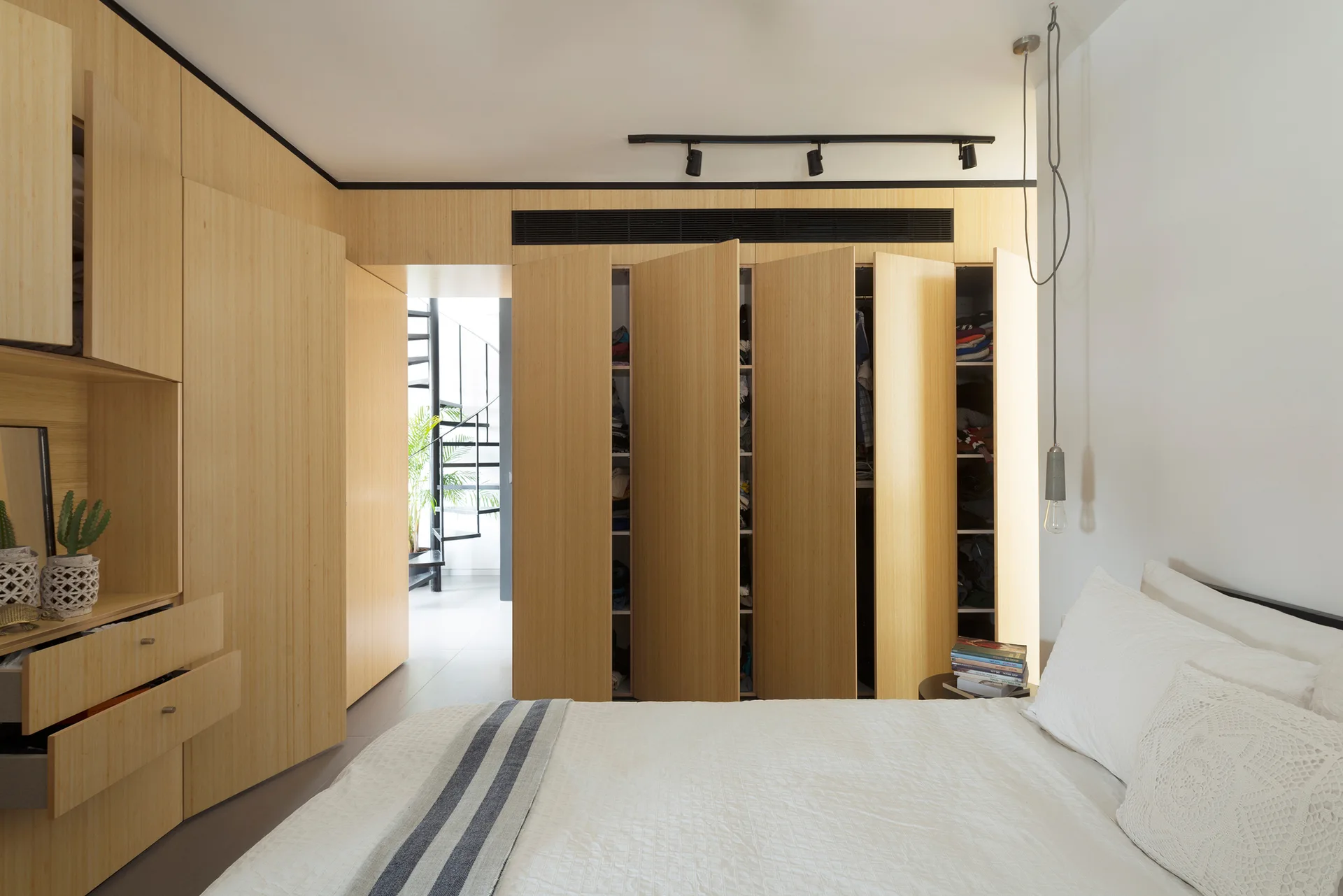 A modern bedroom features white bedding and a wall of built-in light wood wardrobes, with an open doorway revealing a black spiral staircase.