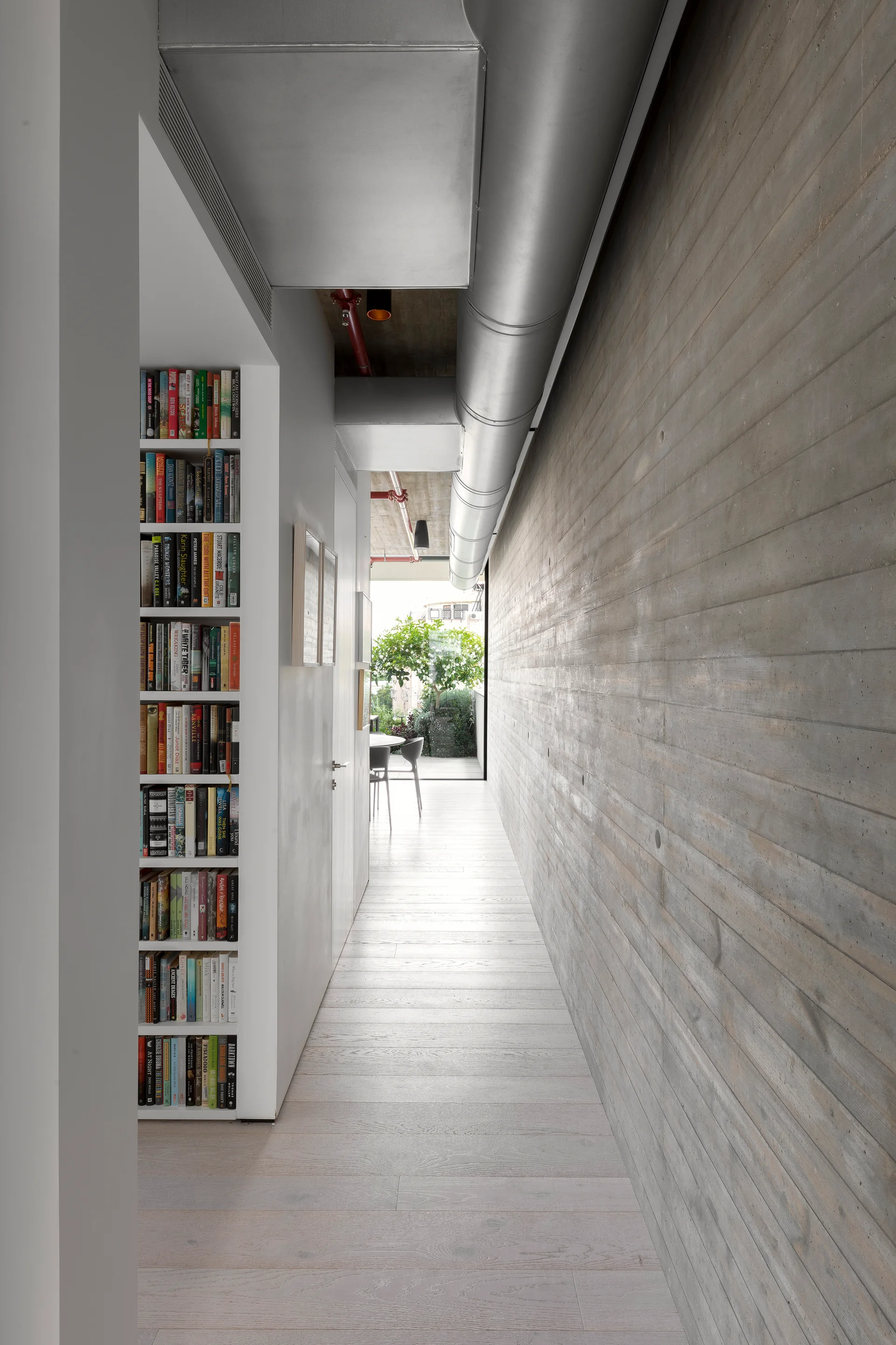 A long hallway features a board-formed concrete wall on one side and a recessed vertical bookcase built into the opposing white wall.