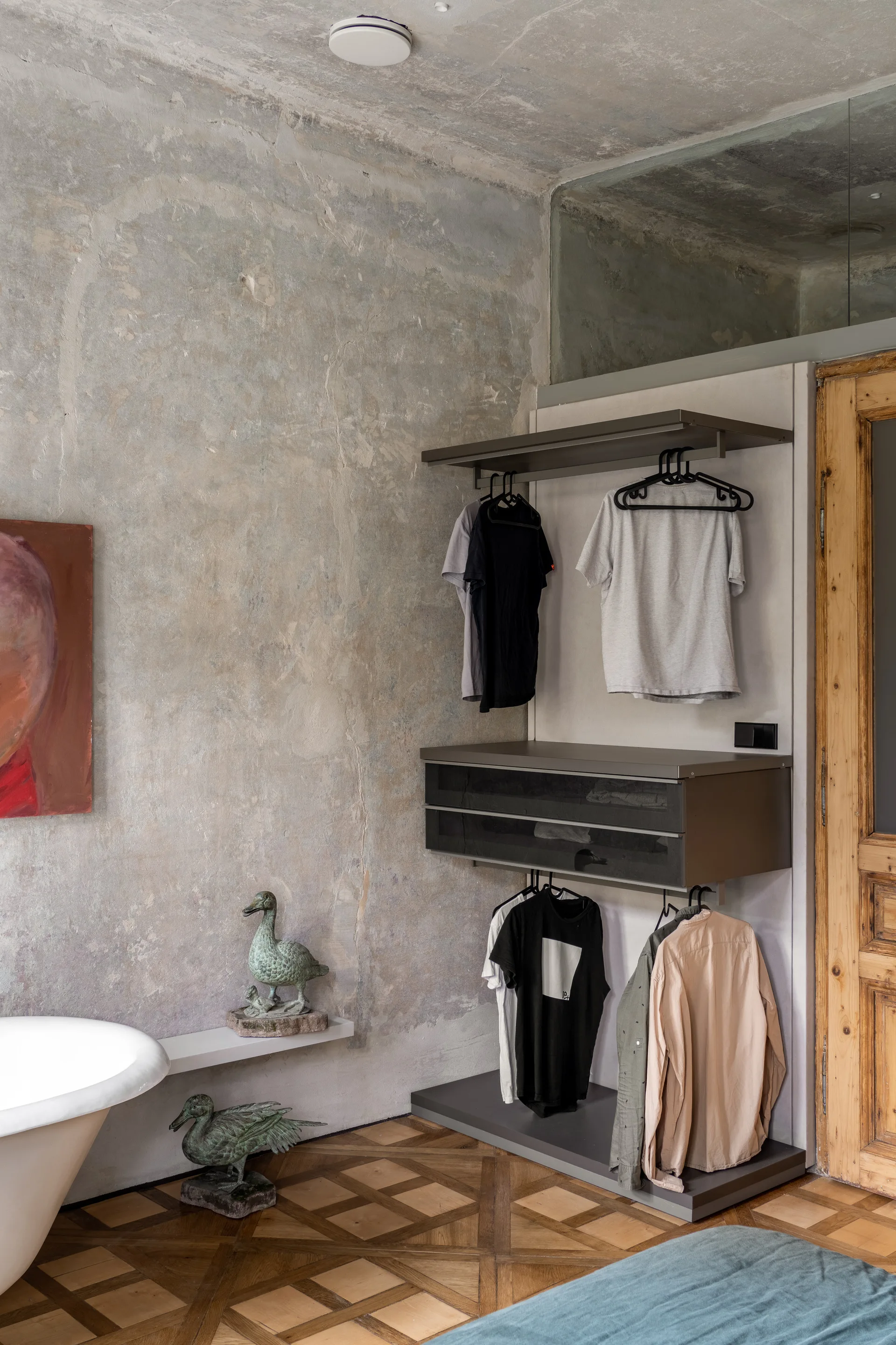 An open metal wardrobe system with hanging clothes sits against a textured concrete wall next to a patterned parquet wood floor.