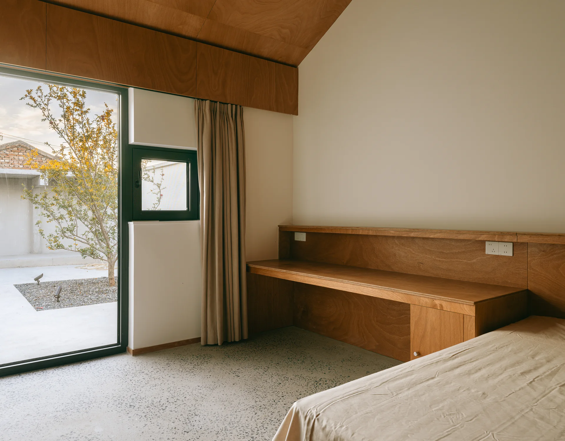 A minimalist bedroom featuring a built-in wooden desk, light terrazzo flooring, and a glass door that opens to an outdoor courtyard.
