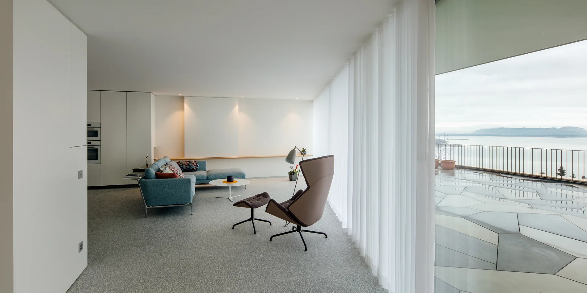 A minimalist living room features a blue sofa, a brown lounge chair with an ottoman, and sheer white curtains along a large window wall.