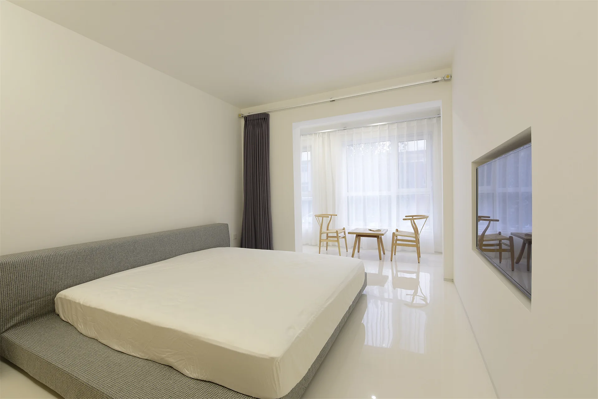 A minimalist white bedroom featuring a low-profile gray fabric bed and two light wood chairs arranged in a sunlit window nook.